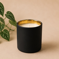 A matte black candle jar with a gold interior and white wax, styled on a beige background next to green trailing plant leaves.