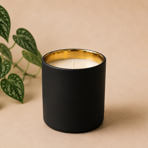 A matte black candle jar with a gold interior and white wax, styled on a beige background next to green trailing plant leaves.