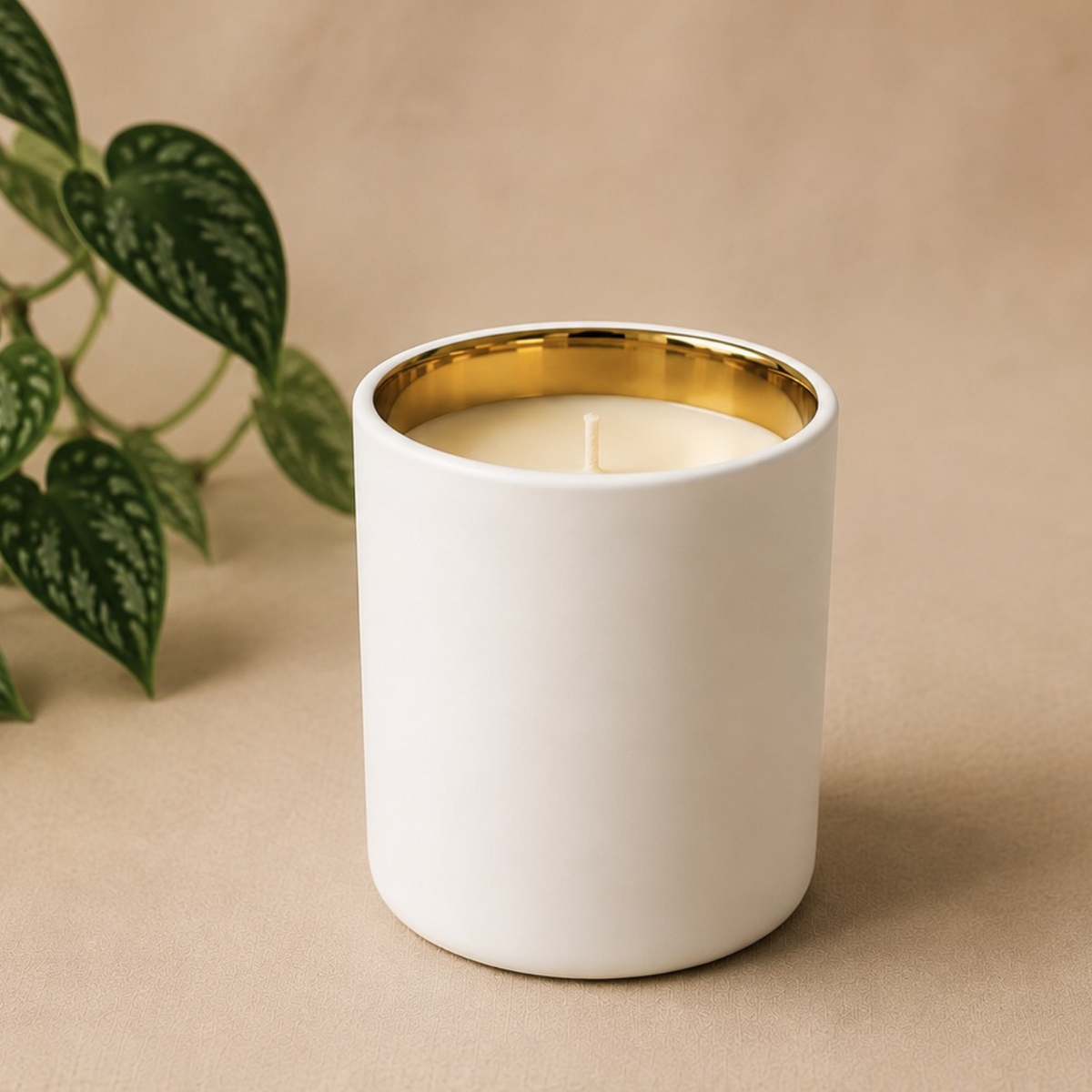 A white ceramic candle jar with a gold inner rim and cream-colored wax, styled on a beige background with green plant leaves in the corner.